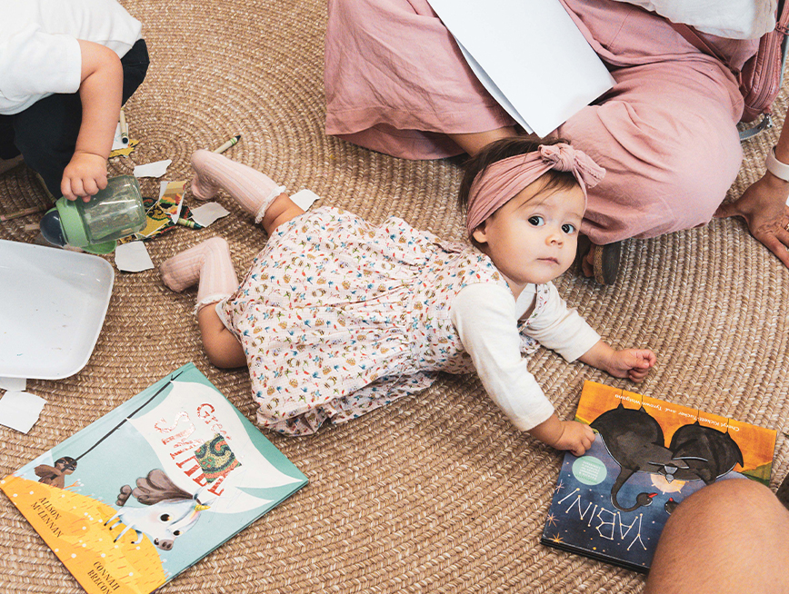 Toddler girl wearing a floral dress, laying down amongst picture books, looking at the camera.