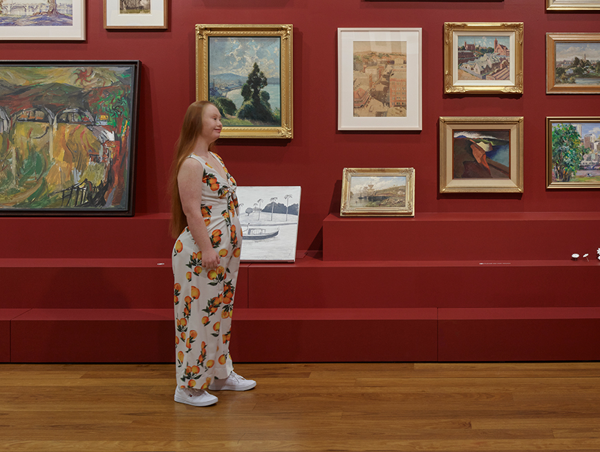 A girl with red hair is wearing a white jumpsuit with lemons all over it and facing sideways. Behind her, there is a red wall featuring thirteen paintings. Most of them are hung on the wall, though there are 3 sitting on plinths.