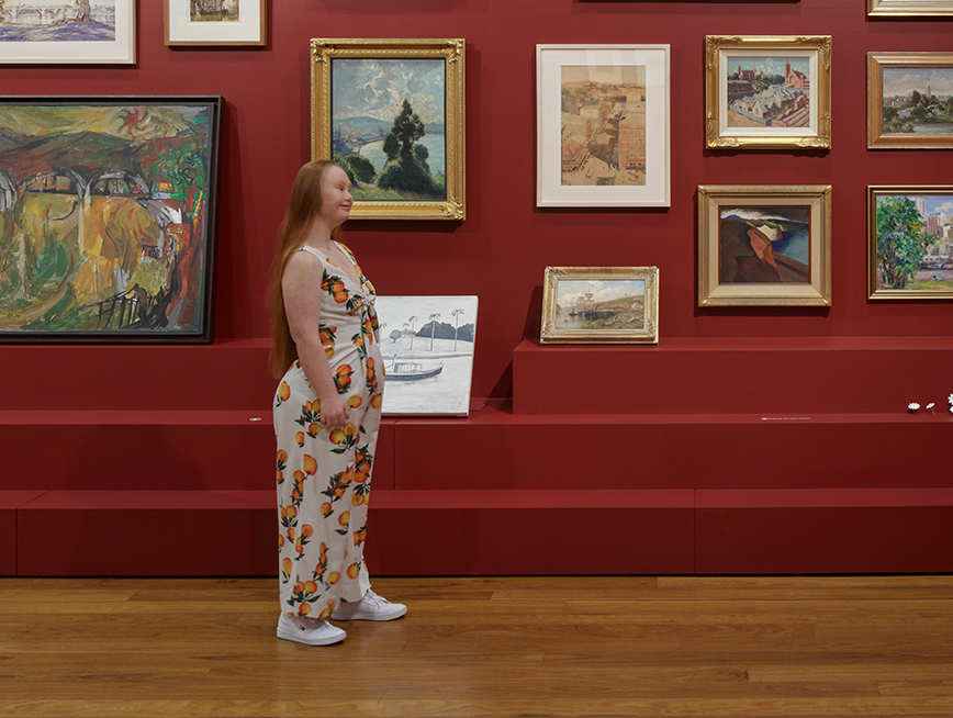 A girl with red hair is wearing a white jumpsuit with lemons all over it and facing sideways. Behind her, there is a red wall featuring thirteen paintings. Most of them are hung on the wall, though there are 3 sitting on plinths.