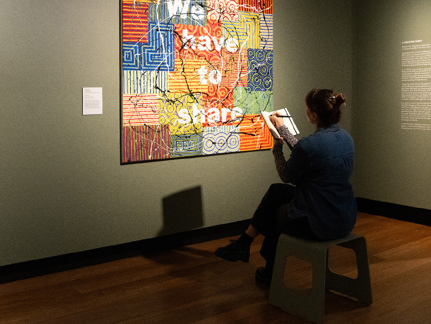 A woman is sitting on a stool at the gallery, writing on her notebook. Facing her, a painting of colourful patchwork with text - 'We have to share' on it.