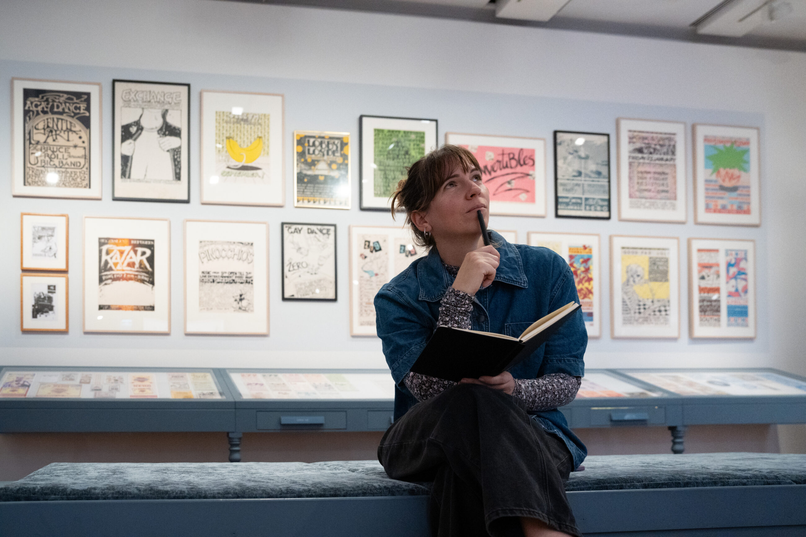 A woman is holding a pen and notebook, sitting on a bench. Behind her are poster art of music events.