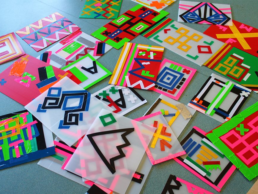 A collection of brightly coloured artworks made from tape and board are scattered all over a grey floor.