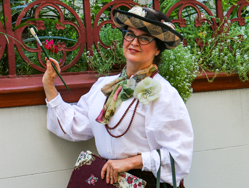 A woman dressed in Victorian-era clothing, wearing glasses, holds a paintbrush in her right hand and a book covered in floral paper in her left hand.