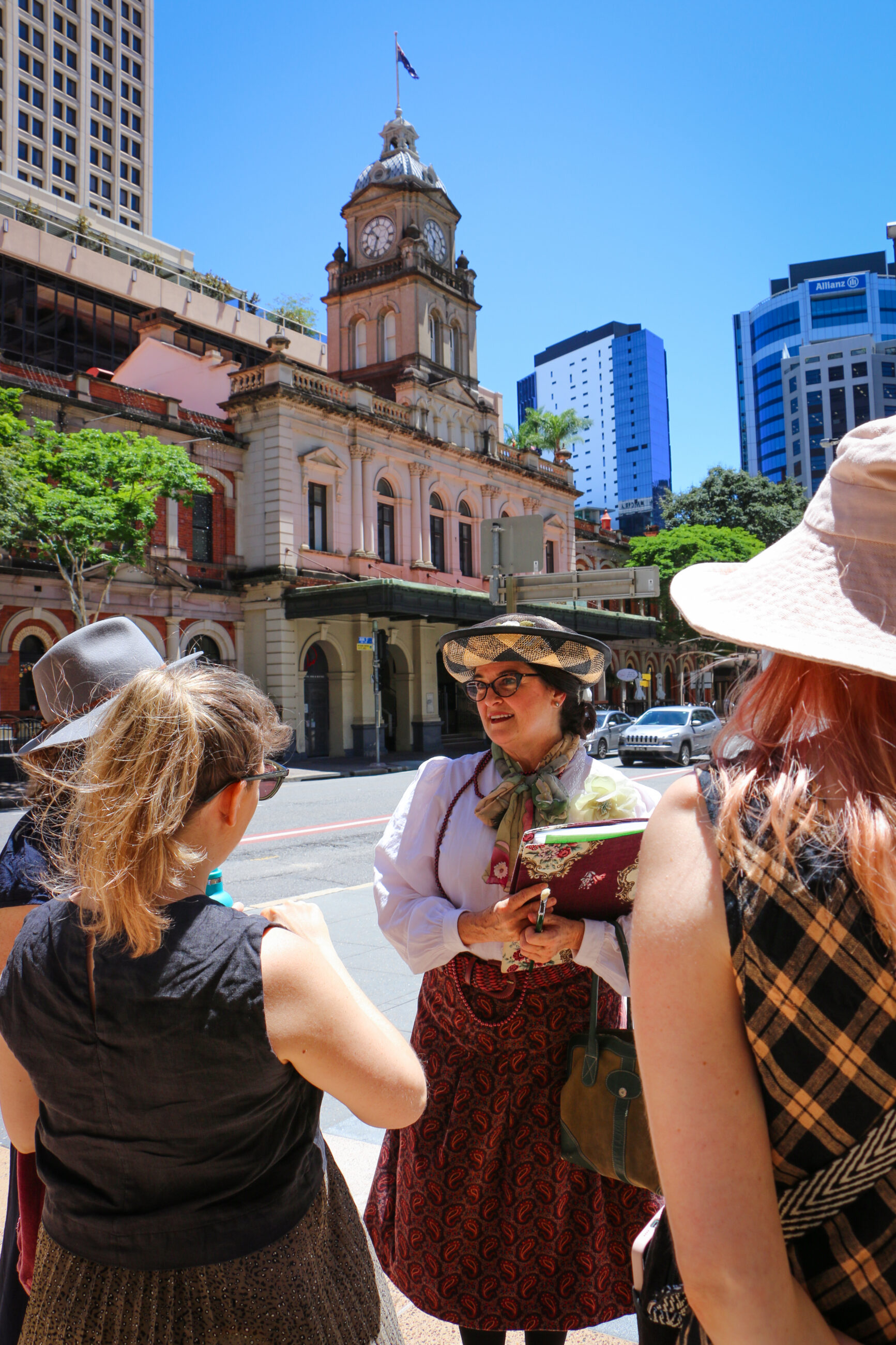 A woman dressed in Victorian-era clothing and wearing glasses holds a paintbrush in her right hand and a book covered in floral paper in her left. She is talking with two other women.
