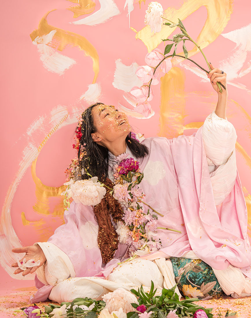 Hiromi Tango sitting in a pink room with white and gold paint over the walls and her face. She's looking up and smiling at a flower that she's holding.