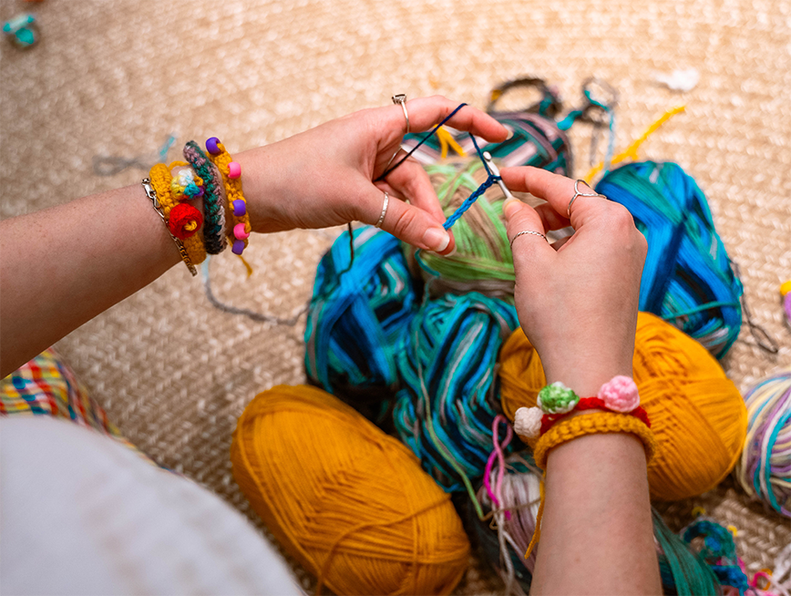 Image of hands crocheting a small chain with colourful yarn in background. On the subjects wrists you can see the friendship bracelets in which are being made.