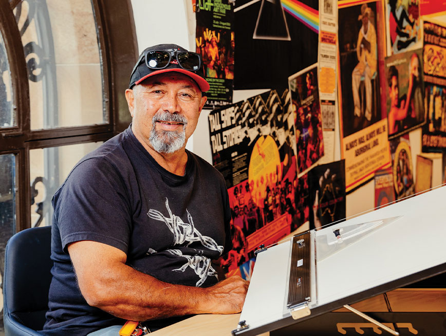 A man wearing a black cap with a red brim sits at a drawing desk and is smiling at the camera. In the background the wall is covered in posters covered in pop culture and political messages.