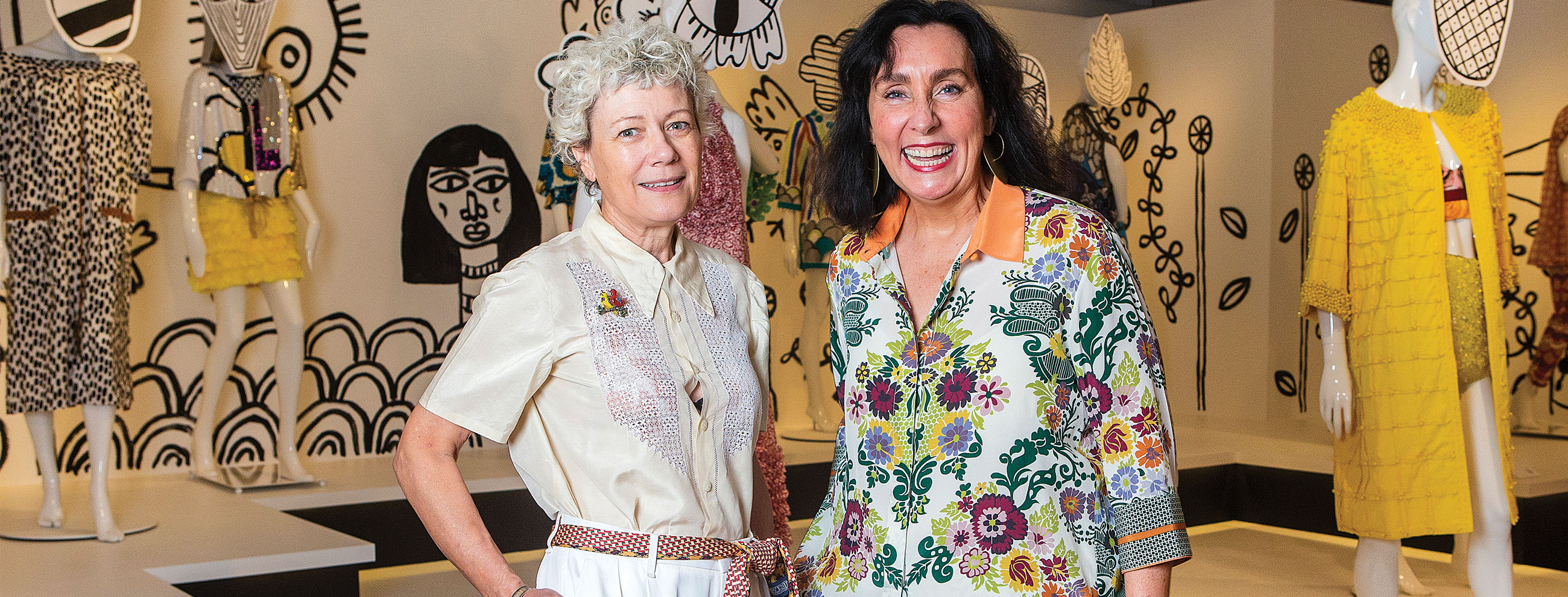 Two women face the camera smiling. They are Lydia Pearson and Pamela Easton, the designers behind Easton Pearson. They are standing in a room surrounding by mannequins wearing bright designs from their own collection.