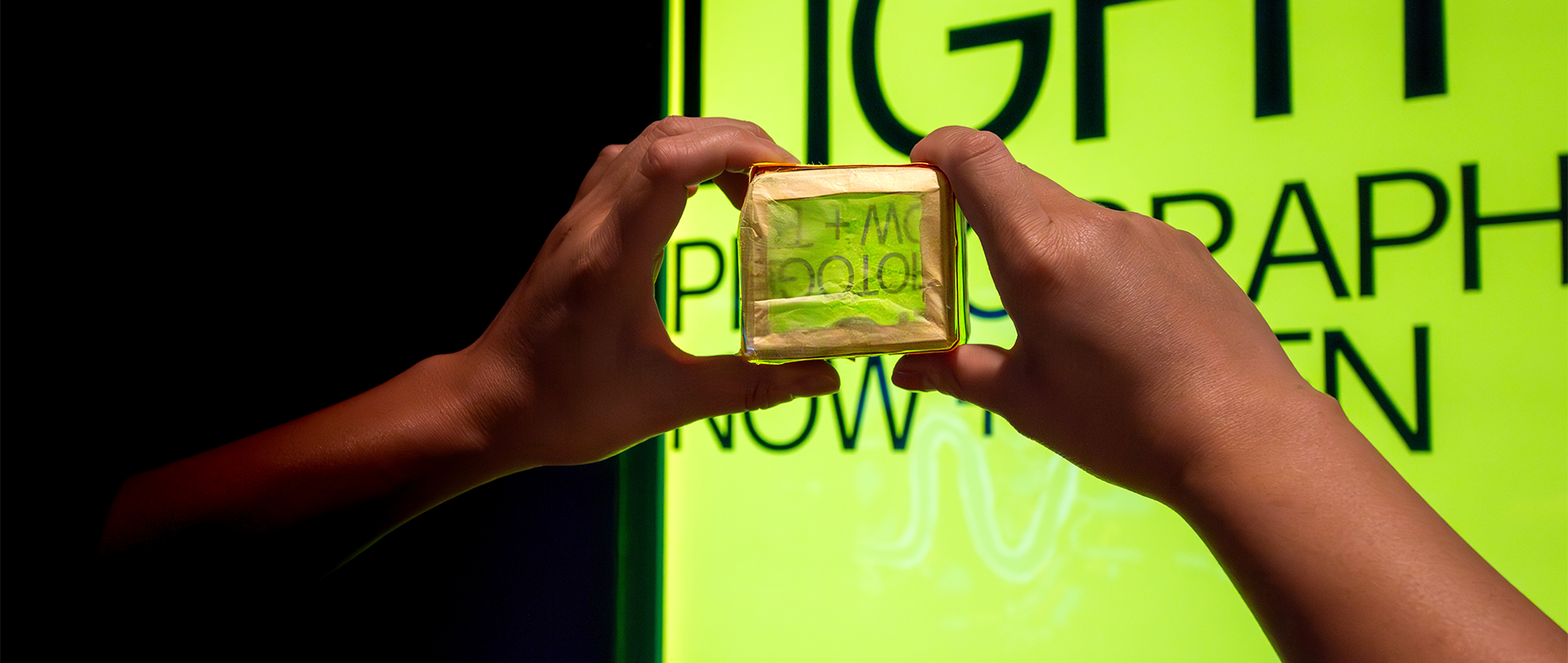 A pair of hands demonstrating Camera Obscura in front of New Light exhibition signage.