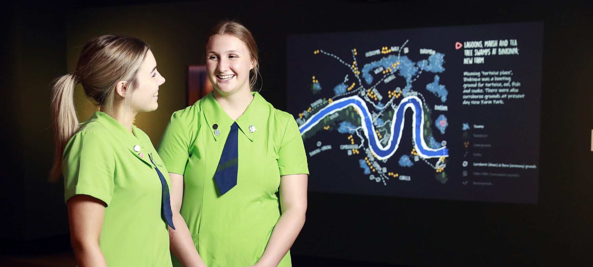 Two students in green uniform standing in front of a display screen.