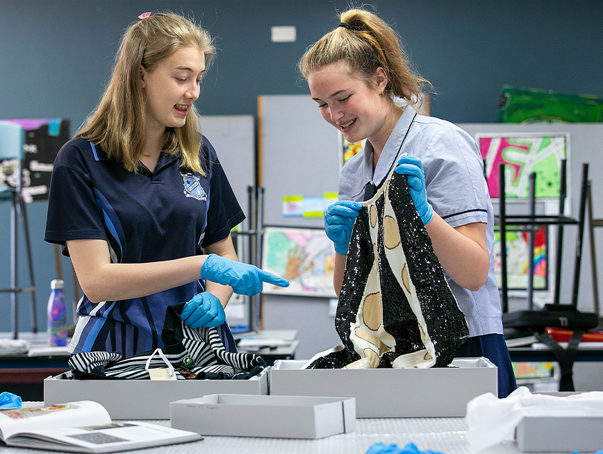 A student wearing blue gloves points to a black and white beaded garment, holding up by another student.