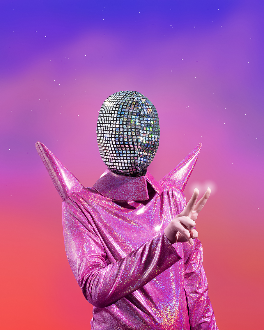 A person in silver mask and pink suit, they hold two fingers up, glowing with light