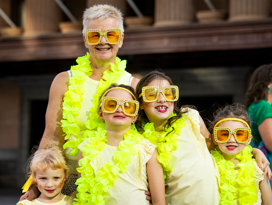 A group of people wearing yellow feathers and yellow thick-framed glasses, smiling at the camera.