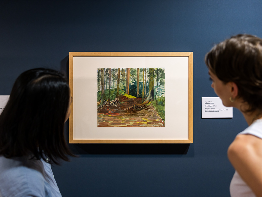 Two people are observing the watercolour painting of a forest scene with green trees and brown earth.