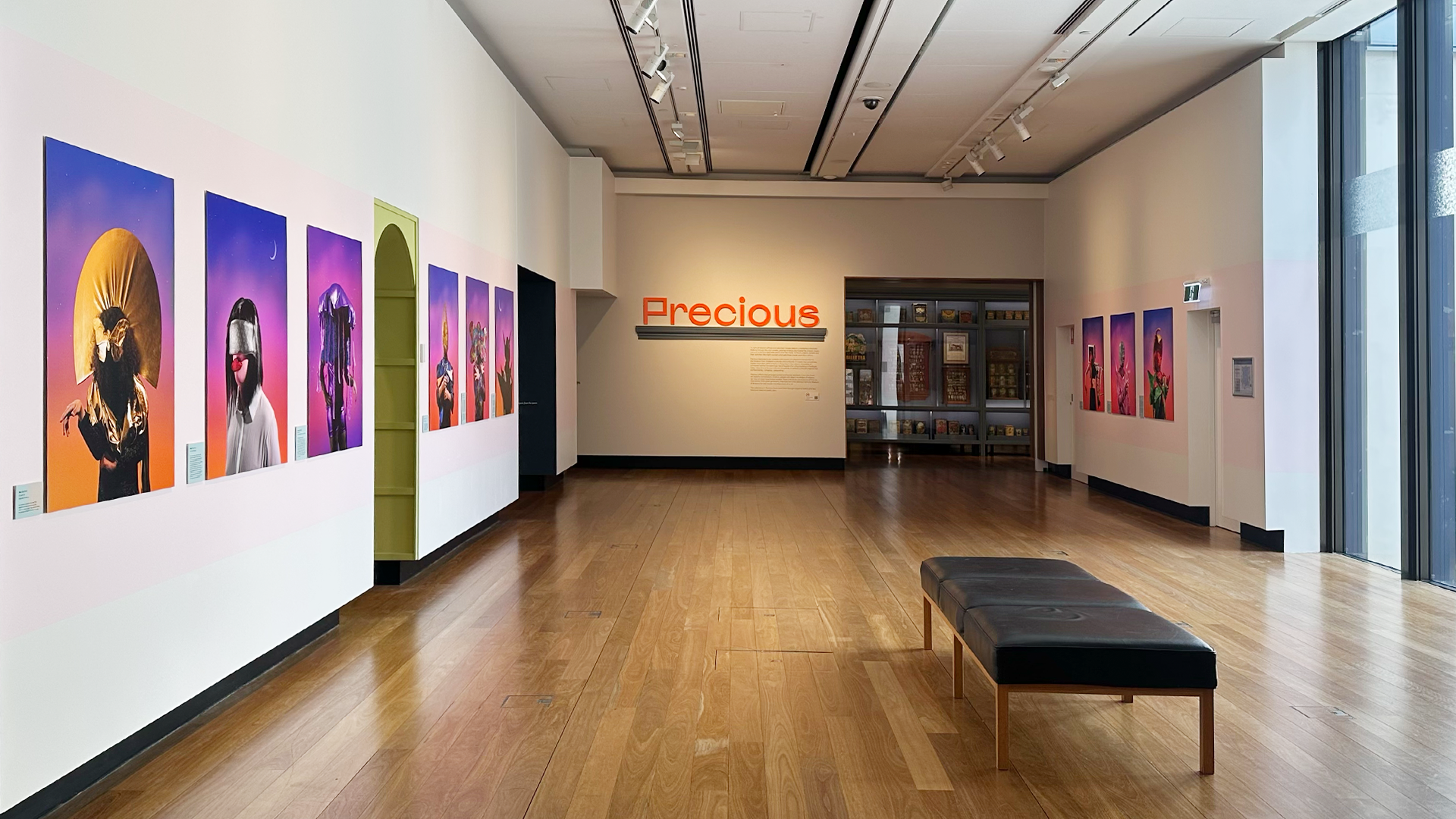 A gallery space with colourful photograph portraits. There is a couch in the middle of the room. Afar, an exhibition entrance with title