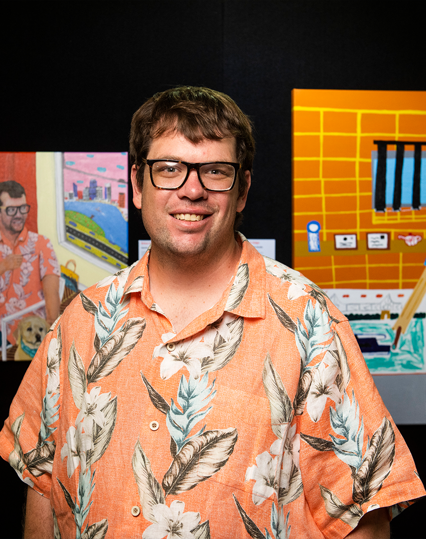 Martin Edge wearing an orange shirt, standing in front of two works of art smiling at the camera.
