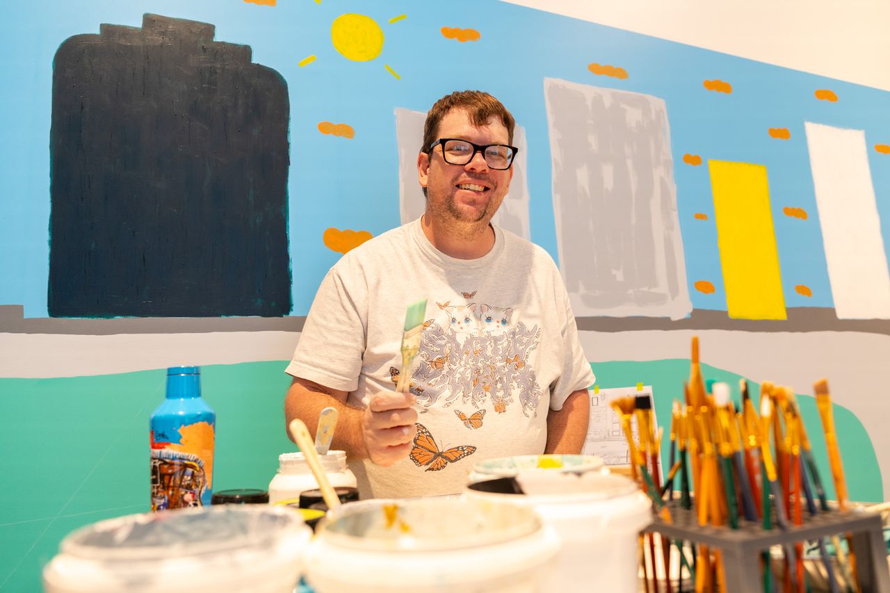 Artist smiling at camera surrounded by painting materials with his mural in the background