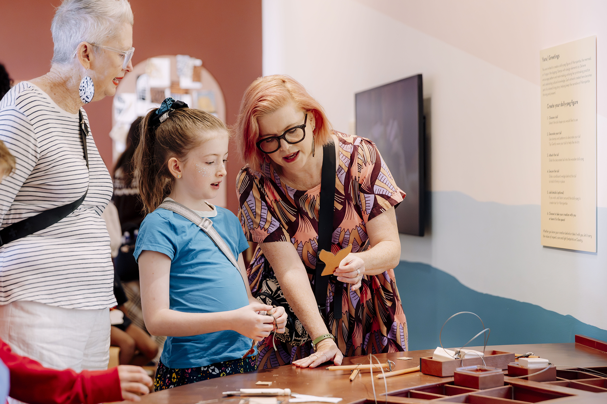 Mum, grandma and a girl at the activity table. mum is showing the girl a mermaid tail.