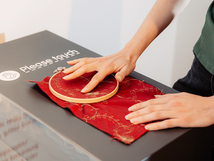 Hands touching the red silk fabric embroidery sample on display.