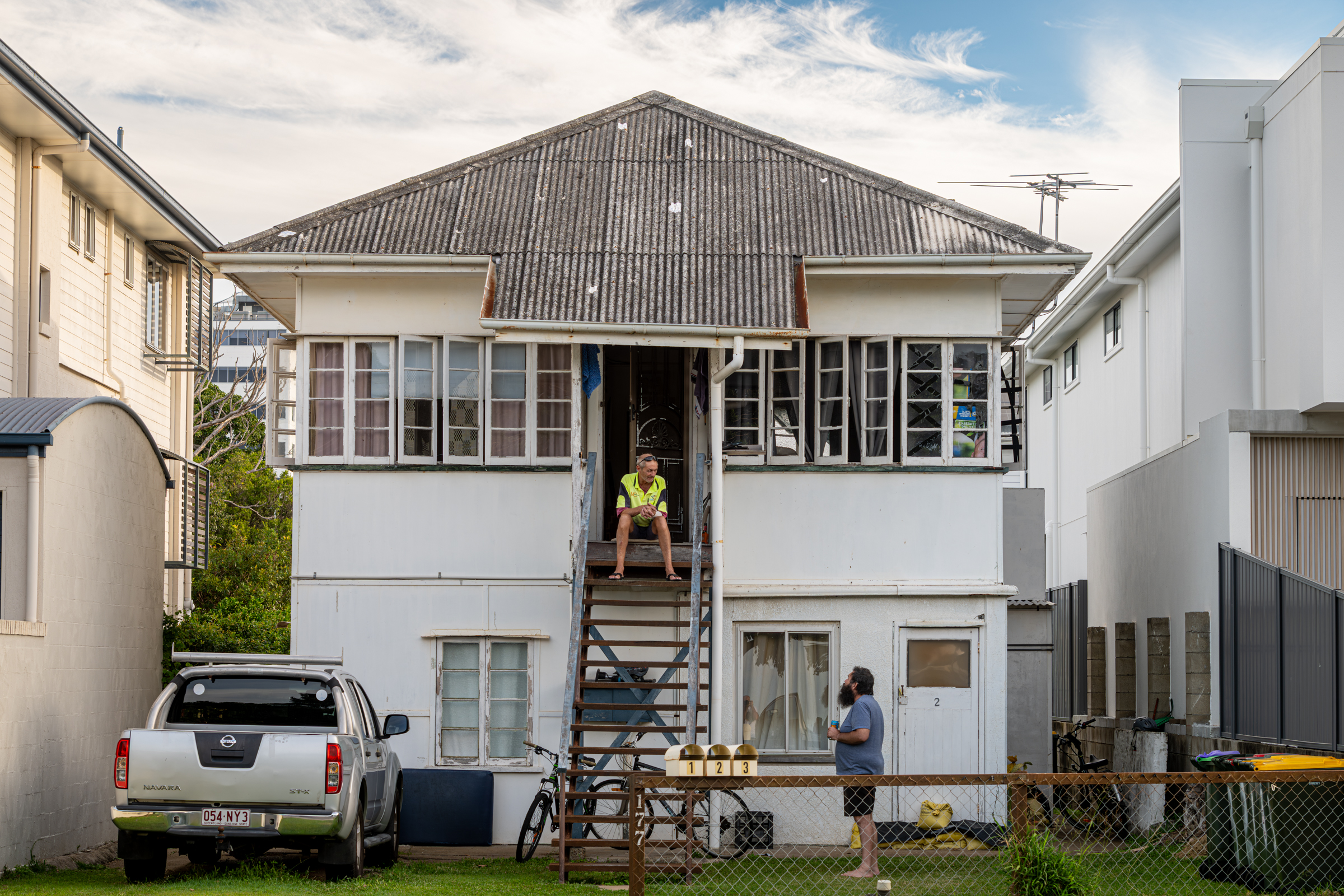 A man sitting on the stairs of a simple Queenslander, having an conversation with another person.
