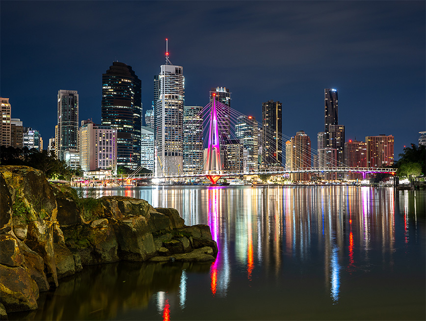 Brisbane city on the horizon with the river below it, reflections and rocks in the image.