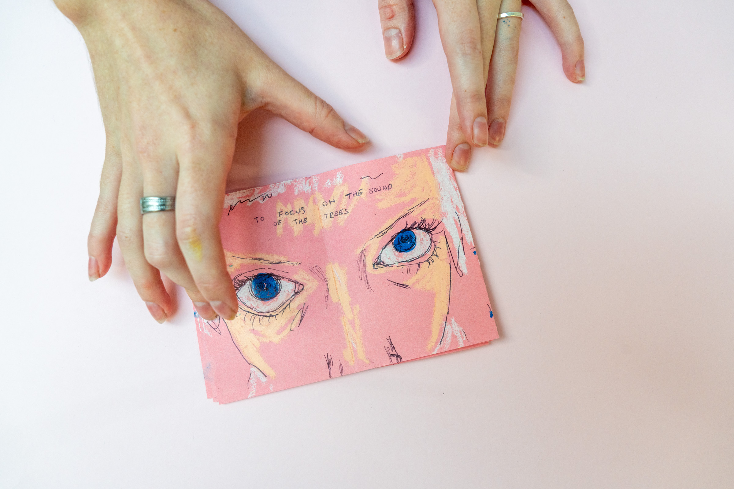 Hands holding open a zine and drawn inside is a woman with blue eyes