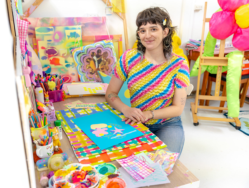 Artist Sunday Jemmott sitting in their studio surrounded by artworks and paper, working on a paper collage on the desk in front of them.