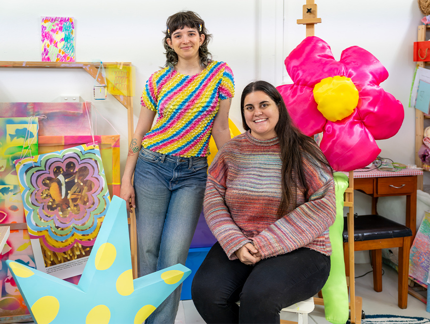 Two women smiling at the camera. One is standing around colourful sculptures.