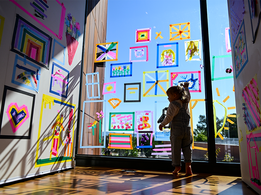 A child taping the windows. They are surrounded by colourful tapes in the gallery.
