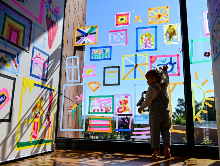 A child taping the windows. They are surrounded by colourful tapes in the gallery.